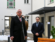 Zbliżenie na starszego mężczyznę w okularach i czarnym płaszczu z czerwonym krawatem, który przemawia do mikrofonu na statywie. Za nim, po prawej stronie, stoi nieco niższy mężczyzna w okularach i ciemnym płaszczu z rękami spllecionymi przed sobą.