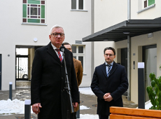 Zbliżenie na starszego mężczyznę w okularach i czarnym płaszczu z czerwonym krawatem, który przemawia do mikrofonu na statywie. Za nim, po prawej stronie, stoi nieco niższy mężczyzna w okularach i ciemnym płaszczu z rękami spllecionymi przed sobą.