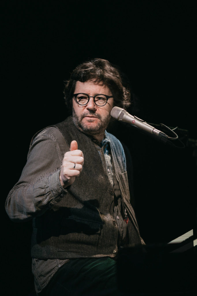 Na zdjęciu artysta siedzący przy fortepianie z mikrofonem przy twarzy.