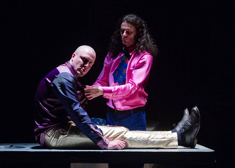 Na zdjęciu dwóch aktorów na czarnym tle. Jeden siedzi z wyprostowanymi nogami, drugi klęczy obok.