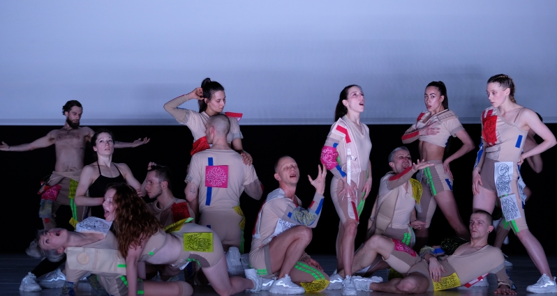 Grupa osób ubranych w cieliste stroje z kolorywami prostokątami. Ustawieni w różnych pozach.