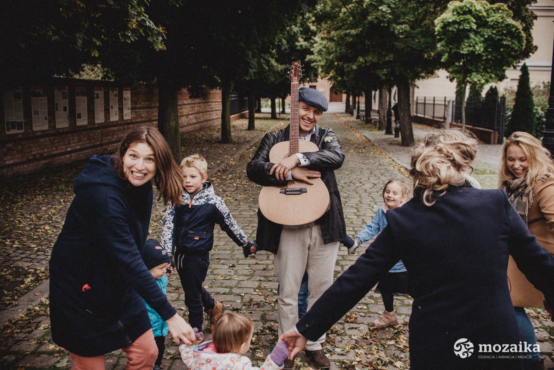 Grupa wesołych kobiet i dzieci tańczy na ulicy trzymając się za dłonie wokół uśmiechniętego mężczyzny przytulającego gitarę. Wzdłuż ulicy rosną drzewa, a w tle widoczne są budynki.