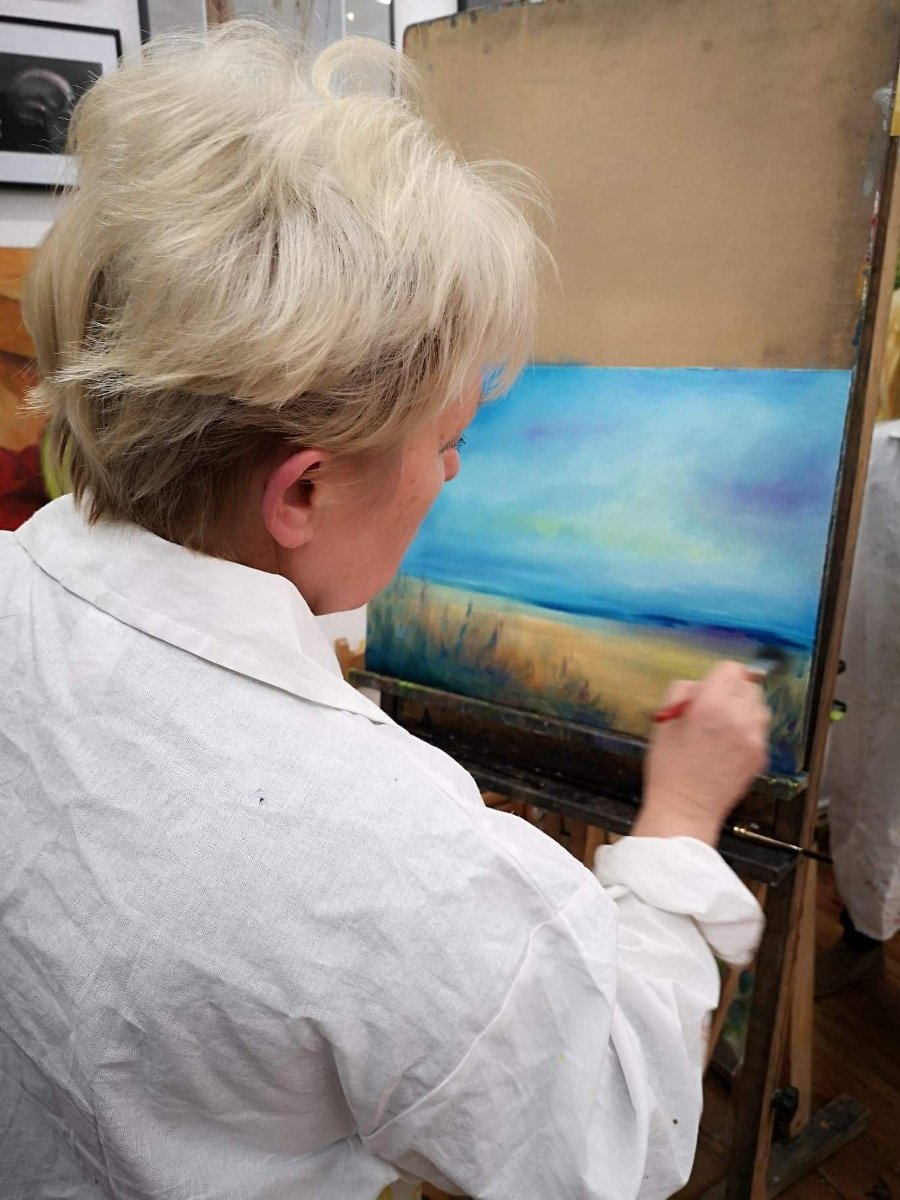 Starsza kobieta w białym fartuchu malująca obraz pędzlem na płótnie.