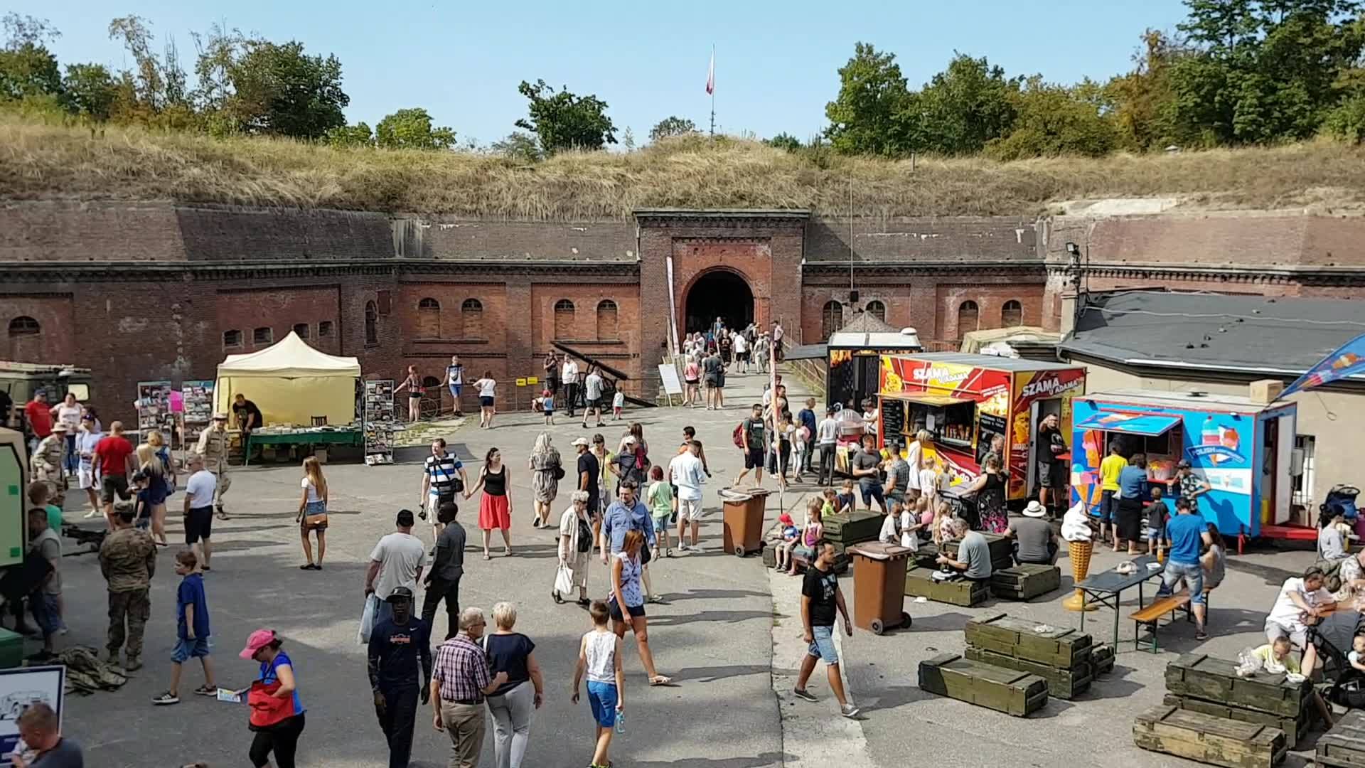Tłumy ludzi w letnich strojach chodzących na dziedzincu Fortu VI. Na nim znajduja się zstragany z jedzeniem, pamiątkami i zabawkami.