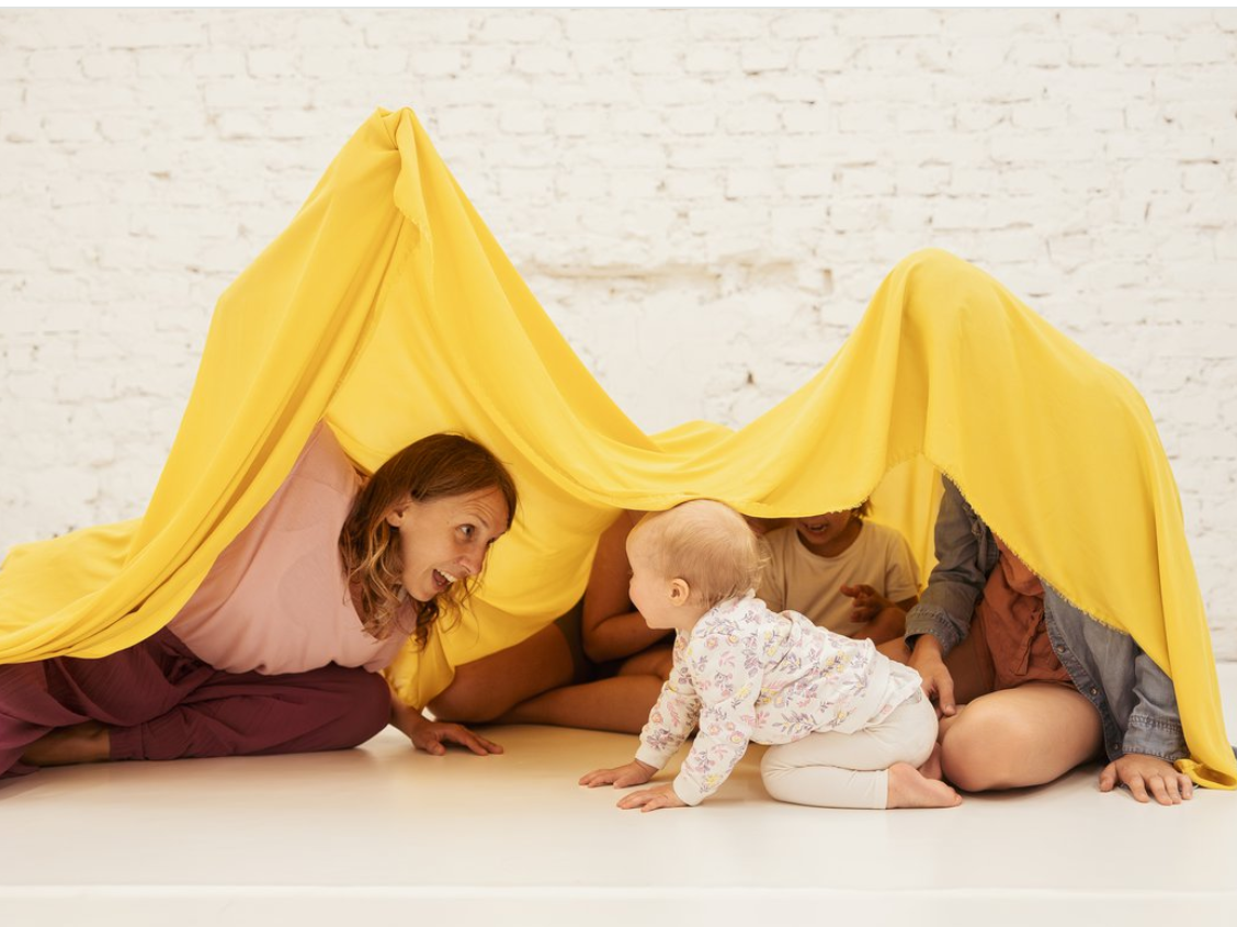 Na zdjęciu fragment warsztatów dla dzieci. Kobieta wraz z kilkorgiem dzieci siedzi na podłodze nakrywając ich żółtym materiałem. Wszyscy są uśmiechnięci.