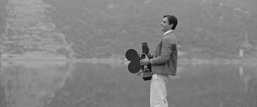 Czarno-białe zdjęcie mężczyzny stojacego bokiem do obiektywu i trzymającego kamerę na tle zamglonego krajobrazu.