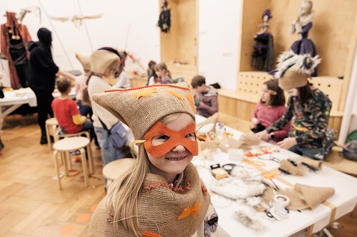 Uśmiechnięta dziewczynka w przebraniu z maską na twarzy. W tle dzieci siedzące przy stole przy pracach plastycznych.