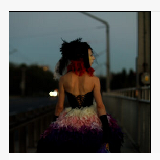 na zdjęciu widoczna postać kobiety idąca wieczorem chodnikiem ulicy, ubrana jest w sukienkę z gorsetem, z odsłoniętymi ramionami, na głowie ma woalkę z czerwonymi i czarnymi piórami i wstążkami.