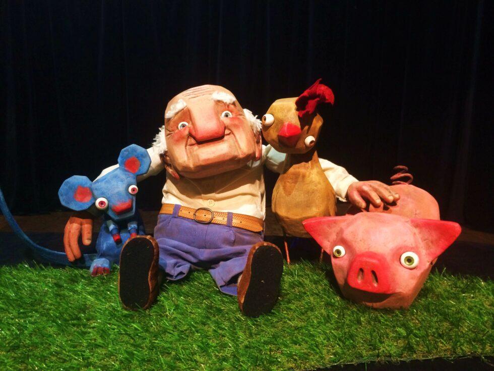 Lalka przedstawiająca starszego mężczyzny obejmującego myszkę, koguta i świnkę. Czarne tło.