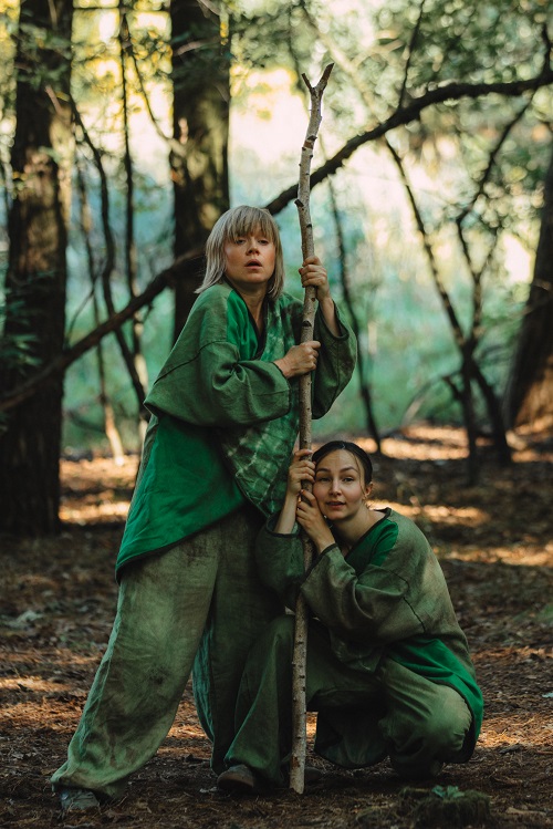 Dwie kobiety w lesie, ubrane w zielone atroje, trzymają cienki pień drzewa.