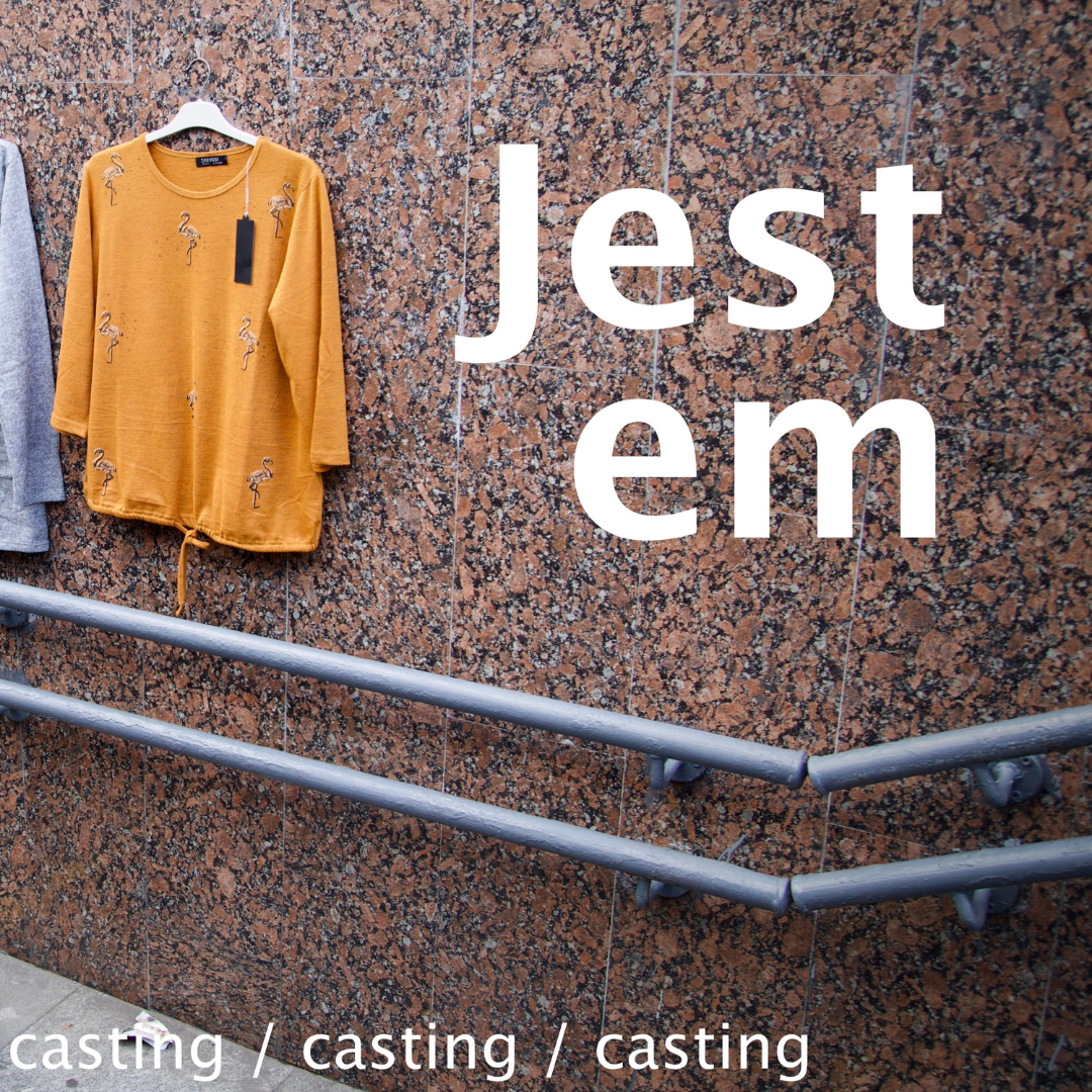 Fragment ściany z poręczą, na ścianie wisi pomarańczowa bluzka z metką od ceny. Na środku napis w dwóch linijkach "jest em".