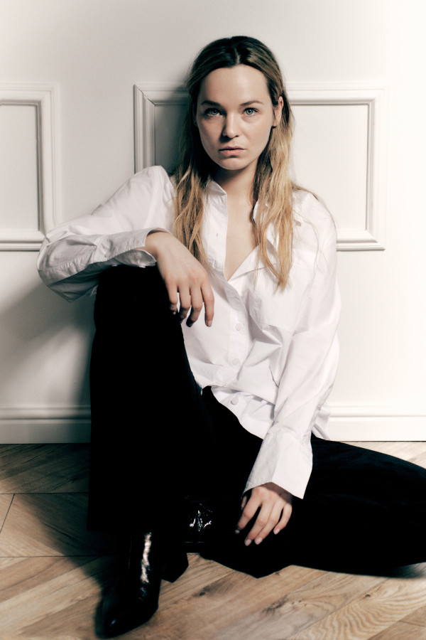 Zdjęcie Michaliny łabacz - młodej kobiety, w białej bluzce i czarnych spodniach, siedzacej pod ścianą, z jedną nogą zgiętą w kolanie.