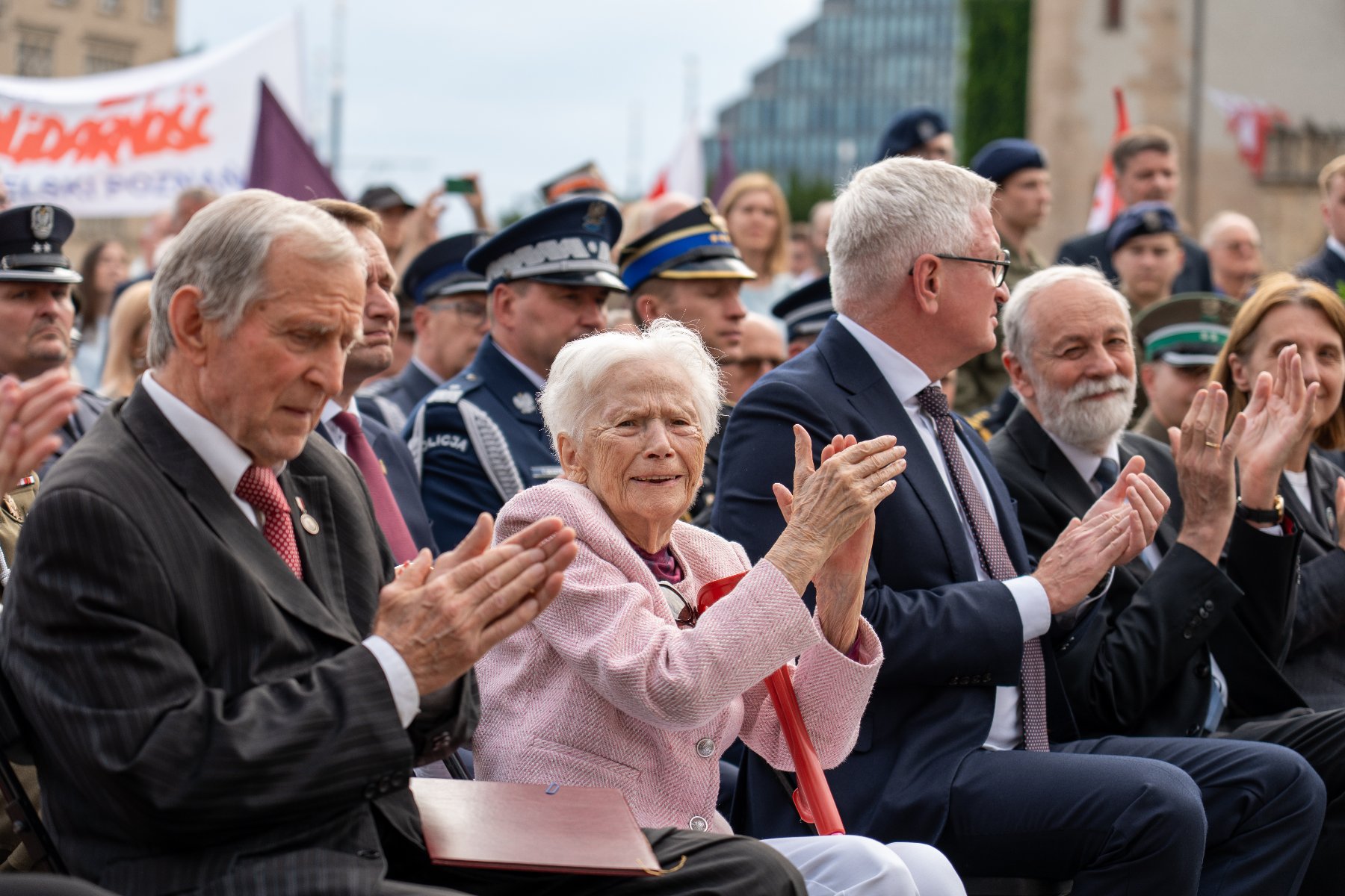 Zdjęcie przedstawia Aleksandrę Banasiak, siedzącą wśród innych ludzi podczas obchodów Poznańskiego Czerwca 1956 r. Starsza kobieta klaszczę i uśmiecha się do aparatu.