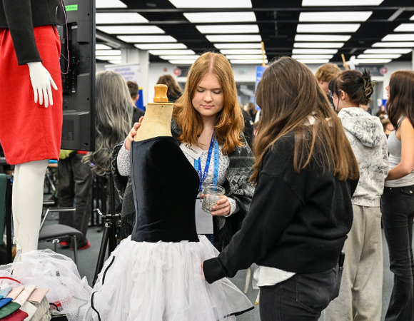 Zdjęcie przedstawia dwie młode dziewczyny ubierające manekina. W tle widać innych młodych ludzi.