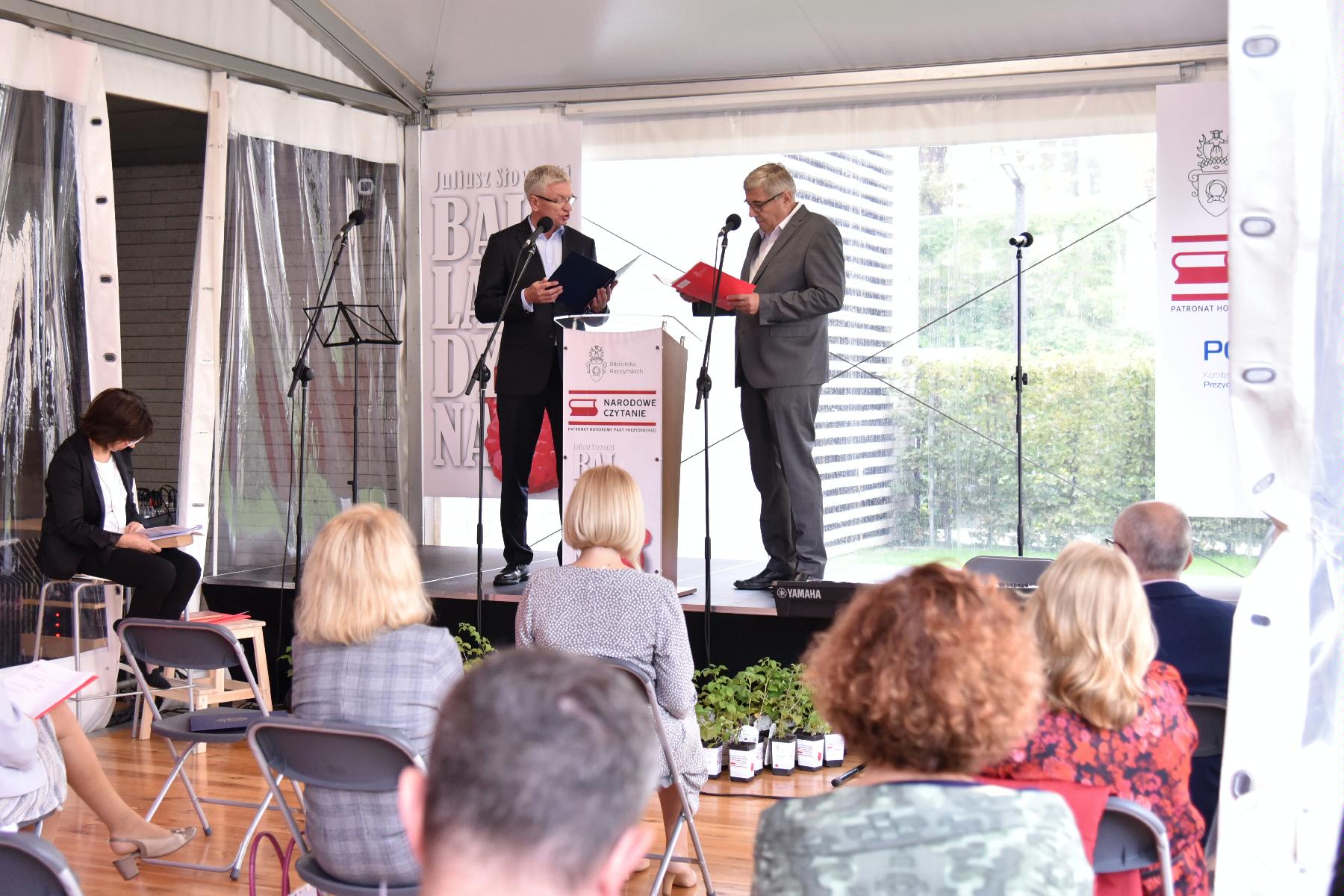 Zdjęcie przedstawia dwóch mężczyzn na scenie i ludzi siedzących na krzesłach pod sceną.