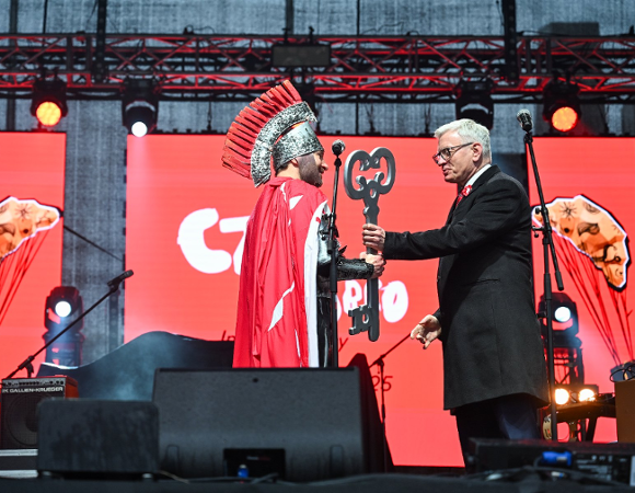Zdjęcie przedstawia dwóch mężczyzn na scenie podczas przekazania kluczy. Jeden w przebraniu rycerza.