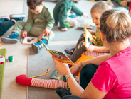 Zdjęcie przedstawia dzieci i panią opiekunkę siedzących na dywanie i czytających książeczki.