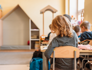 Zdjęcie przedstawia dzieci siedzące w ławkach tyłem do aparatu.