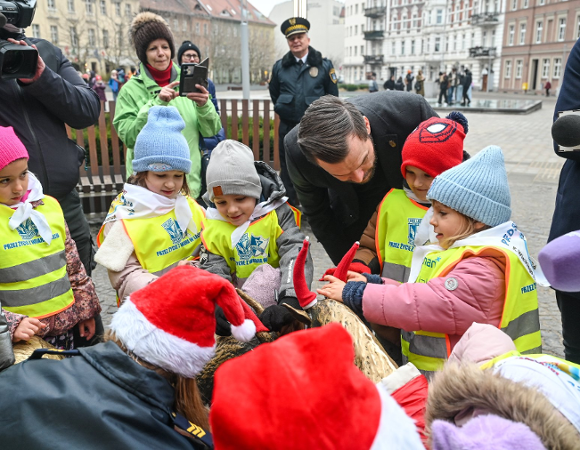 Zdjęcie przedstawia dzieci ubierające koziołki i stojących obok nich dorosłych, w tym dziennikarzy z mikrofonami i kamerami.