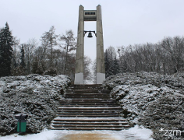 Zdjęcie przedstawia dzwon i pomnik, na którym jest umieszczony oraz schody i zieleń - krzewy i drzewa pokryte śniegiem.