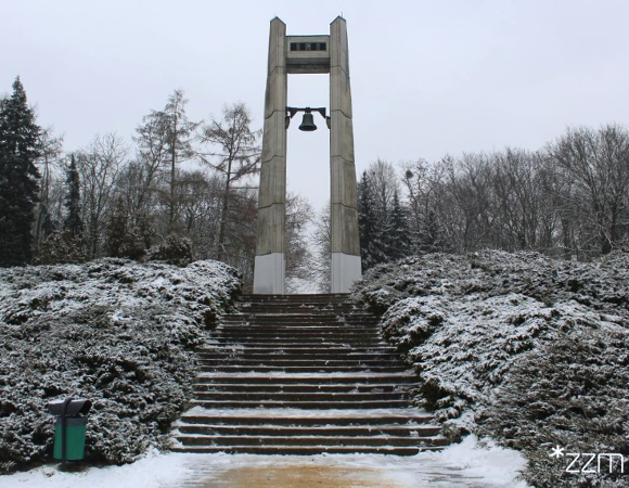 Zdjęcie przedstawia dzwon i pomnik, na którym jest umieszczony oraz schody i zieleń - krzewy i drzewa pokryte śniegiem.