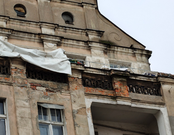 Zdjęcie przedstawia fragment silnie zniszczonej elewacji zabytkowej kamienicy, na której widoczne są głębokie ubytki tynku odsłaniające gołą cegłę oraz uszkodzone detale architektoniczne, takie jak ozdobne tralki balkonu.