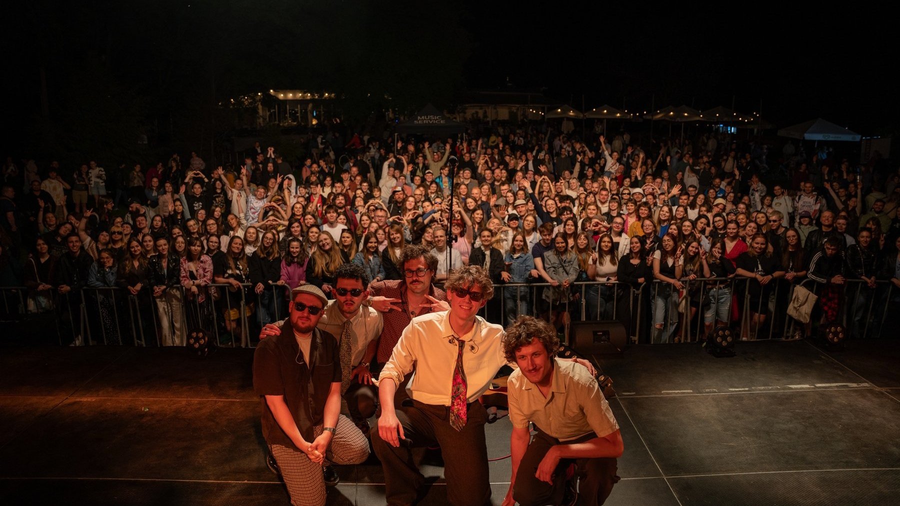 Zdjęcie przedstawia grupę artystów pozujących do zdjęcia. W tle, za barierkami, wiwatujący tłum.