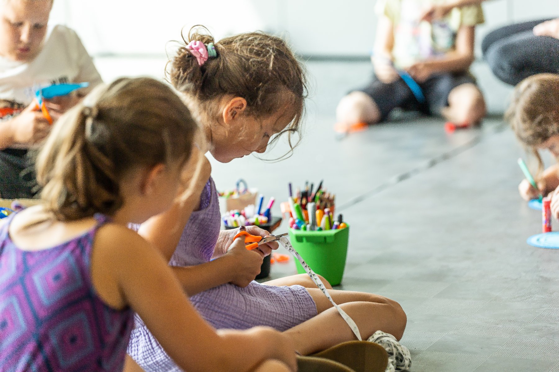 Zdjęcie przedstawia grupę dzieci, siedzących na podłodze. Niektóre rysują, inne wycinają.
