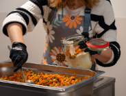 Zdjęcie przedstawia kobietę nakładającą jedzenie do słoika. W misce przed nią znajdują się pokrojony groszek i marchewka.