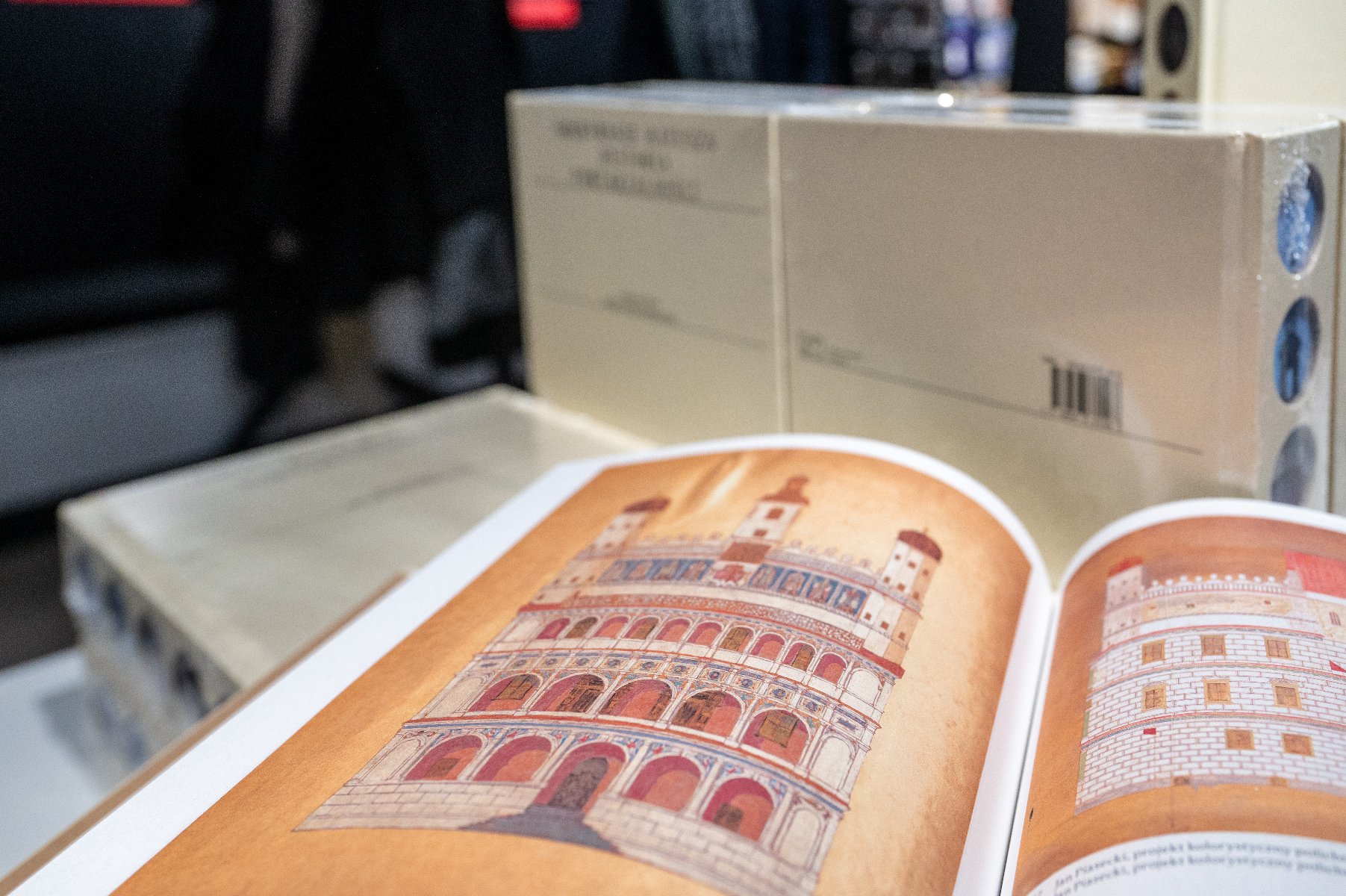 Zdjęcie przedstawia kobietę podpisującą książkę przy stoliku. Za nią widać innych ludzi.