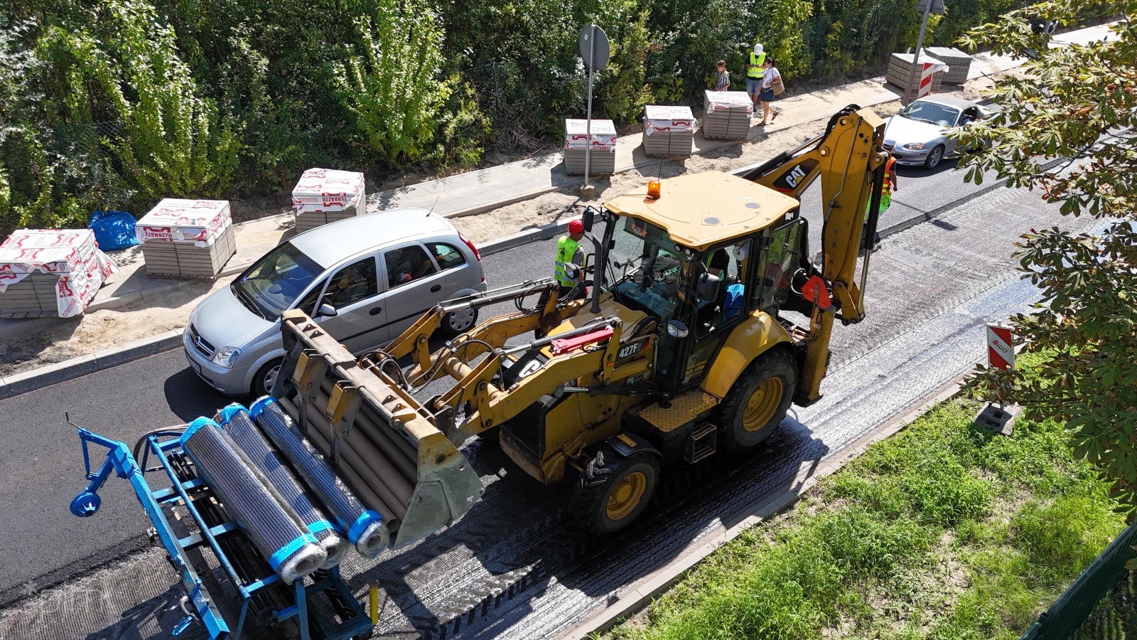 Zdjęcie przedstawia maszynę budowlaną pracującą na jezdni. Obok widać robotników.