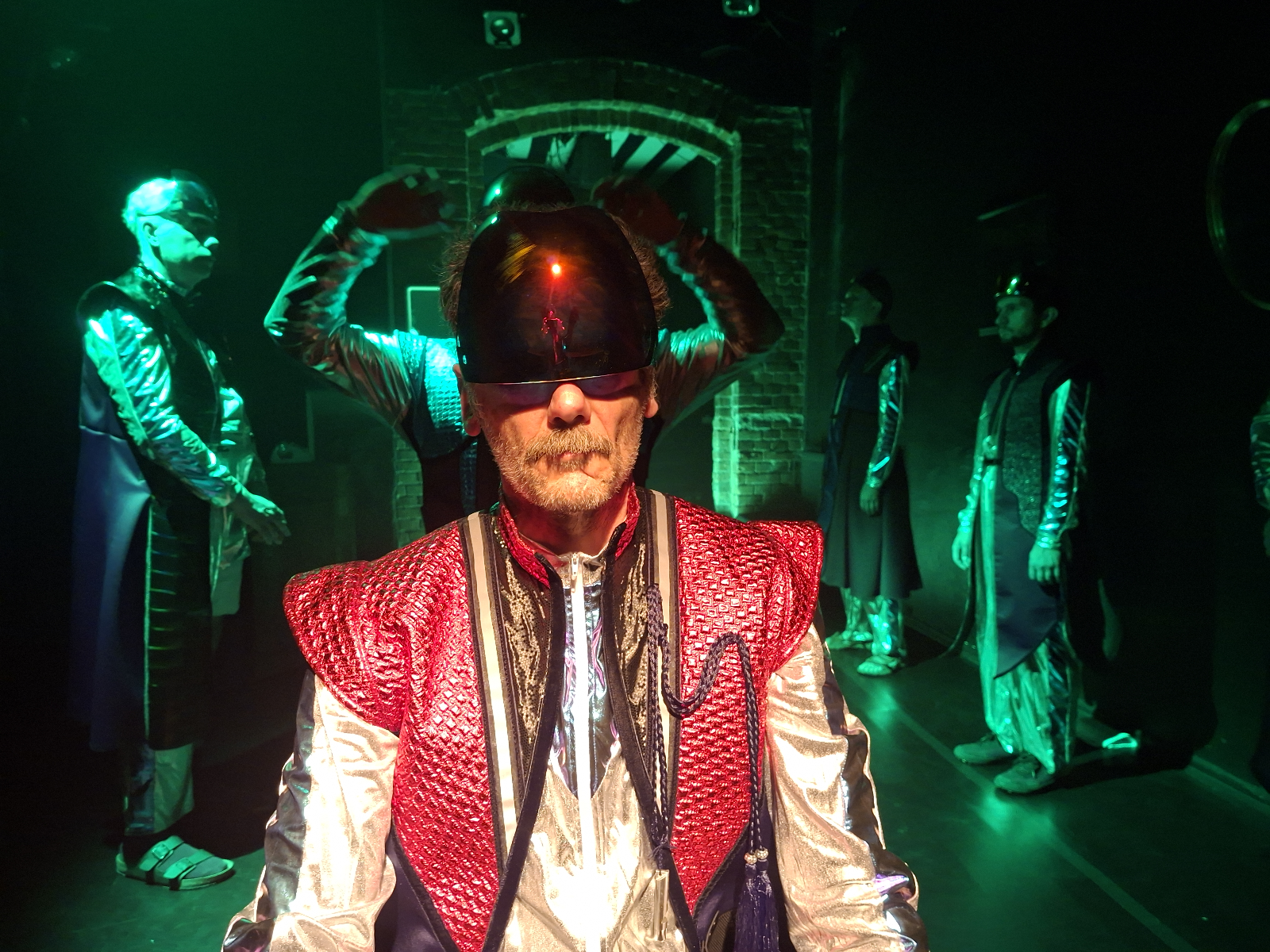 Zdjęcie przedstawia mężczyznę w średniowiecznym stroju z dziwnym kaskiem na głowie. W tle widać innych mężczyzn.