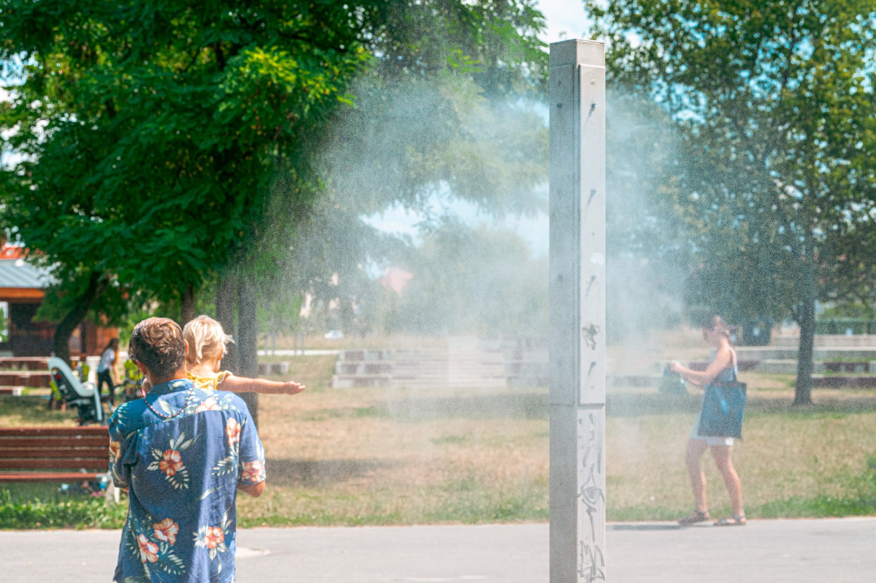 Zdjęcie przedstawia mglawicę wodną wśród zieleni oraz chłodzących się przy niej ludzi. To mężczyzna z dzieckiem na rękach i młoda kobieta.
