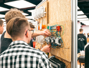 Zdjęcie przedstawia młodych chłopaków naprawiających jakieś urządzenie zawieszone na ścianie.