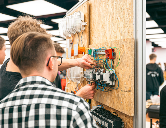 Zdjęcie przedstawia młodych chłopaków naprawiających jakieś urządzenie zawieszone na ścianie.