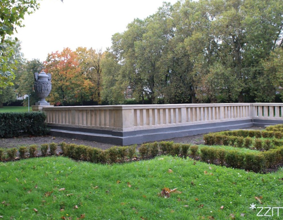 Zdjęcie przedstawia murek i betonową wazę w parku wśród zieleni.