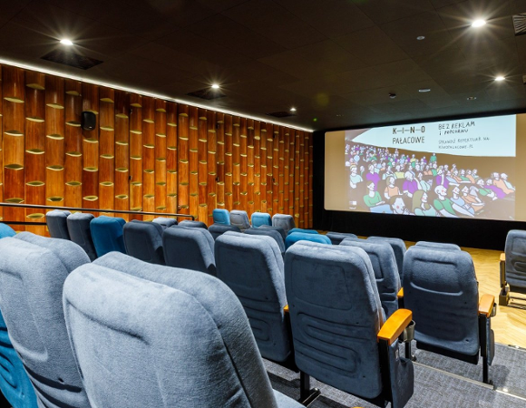 Zdjęcie przedstawia nowoczesną, kameralną salę Kina Pałacowego w Poznaniu, w której znajdują się rzędy wygodnych, niebieskich foteli skierowanych w stronę dużego ekranu projekcyjnego wyświetlającego ilustrowaną grafikę z napisem "K-I-N-O PAŁACOWE - BEZ REKLAM i popcornu".