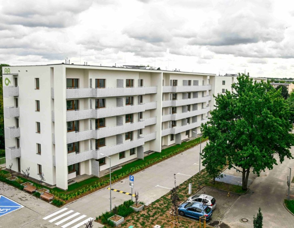 Zdjęcie przedstawia nowoczesny, pięciokondygnacyjny budynek mieszkalny o białej elewacji, wyposażony w liczne balkony z jasnymi, ażurowymi balustradami oraz brązową stolarkę okienną. Przed blokiem znajduje się uporządkowana infrastruktura: asfaltowa droga wewnętrzna, miejsca parkingowe (w tym wyraźnie oznakowane niebieskie miejsce dla osób z niepełnosprawnościami) oraz niska zieleń i młode drzewa.