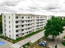 Zdjęcie przedstawia nowoczesny, pięciokondygnacyjny budynek mieszkalny o białej elewacji, wyposażony w liczne balkony z jasnymi, ażurowymi balustradami oraz brązową stolarkę okienną. Przed blokiem znajduje się uporządkowana infrastruktura: asfaltowa droga wewnętrzna, miejsca parkingowe (w tym wyraźnie oznakowane niebieskie miejsce dla osób z niepełnosprawnościami) oraz niska zieleń i młode drzewa.