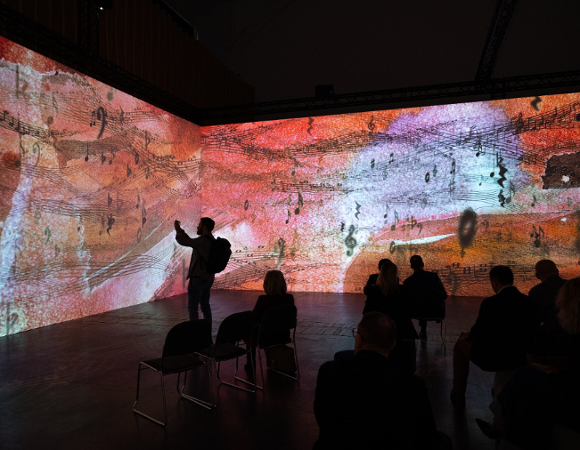 Zdjęcie przedstawia obraz wyświetlany na scianie, przedstawiajacy pięciolinie z nutami, oraz ludzi, którzy go oglądają i robią zdjęcia telefonami.