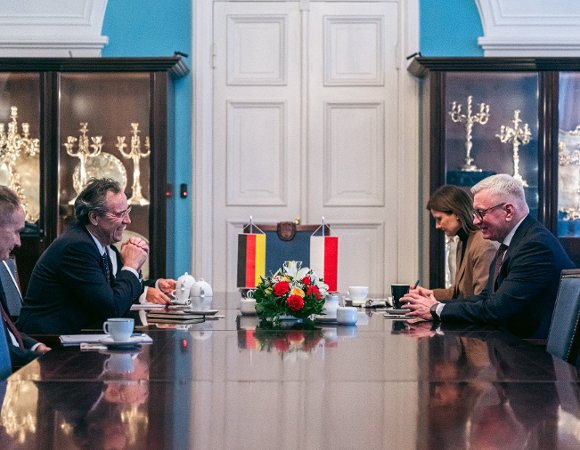 Zdjęcie przedstawia oficjalne spotkanie dwóch delegacji przy długim, lśniącym stole konferencyjnym, na którym ustawiono małe flagi Niemiec i Polski oraz kompozycję z czerwonych i żółtych kwiatów. Po obu stronach stołu siedzą mężczyźni w ciemnych garniturach zaangażowani w rozmowę.