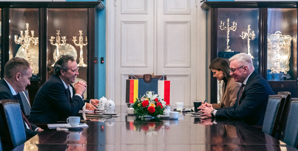 Zdjęcie przedstawia oficjalne spotkanie dwóch delegacji przy długim, lśniącym stole konferencyjnym, na którym ustawiono małe flagi Niemiec i Polski oraz kompozycję z czerwonych i żółtych kwiatów. Po obu stronach stołu siedzą mężczyźni w ciemnych garniturach zaangażowani w rozmowę.