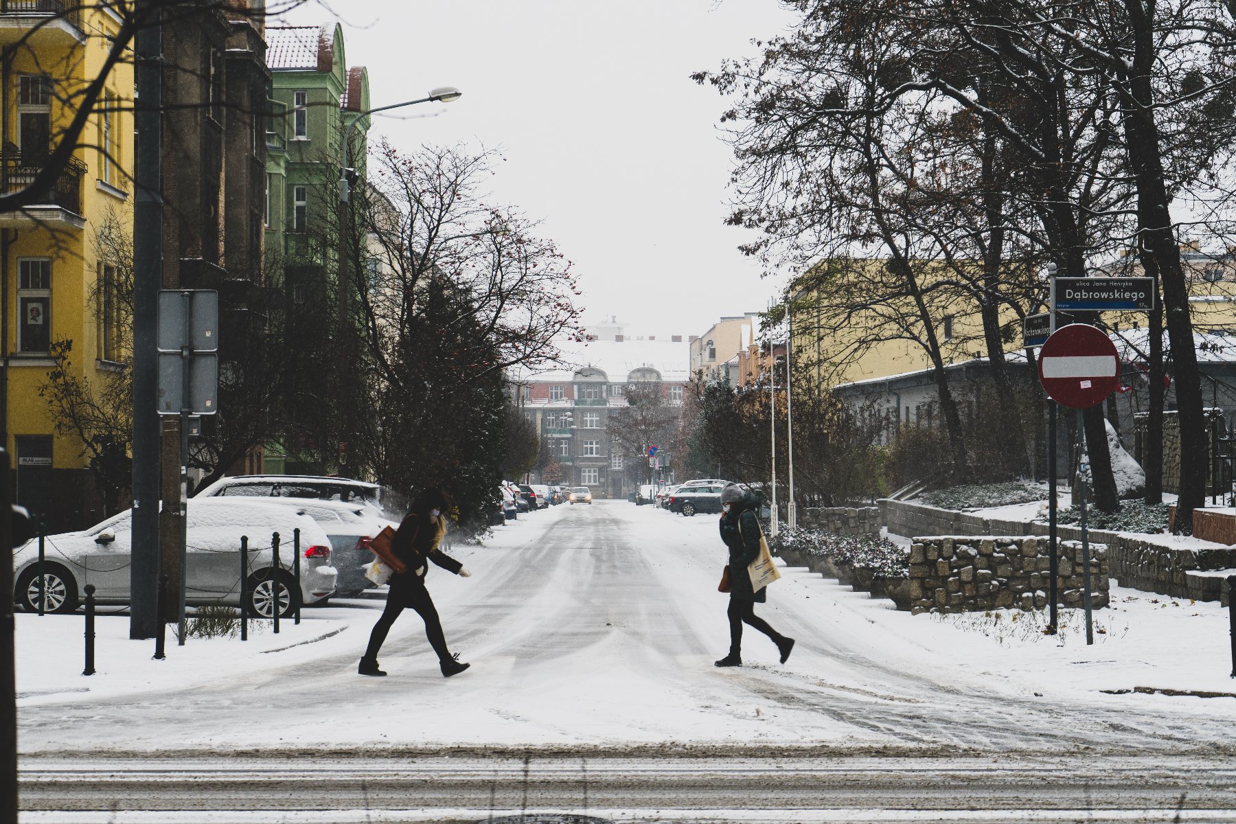 Zdjęcie przedstawia ośnieżoną ulicę, przez którą przechodzą dwie kobiety. Wzdłuż ulicy znajdują się budynki i stoją samochody.