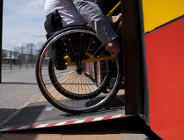 Zdjęcie przedstawia osobę na wózku wjeżdżająca po specjalnej platformie podjazdowej do autobusu.