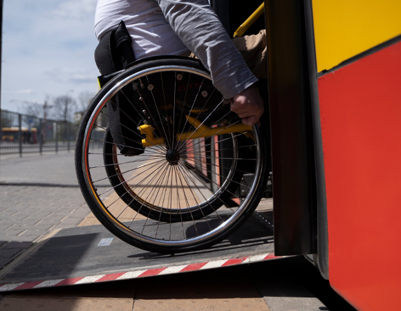 Zdjęcie przedstawia osobę na wózku wjeżdżająca po specjalnej platformie podjazdowej do autobusu.