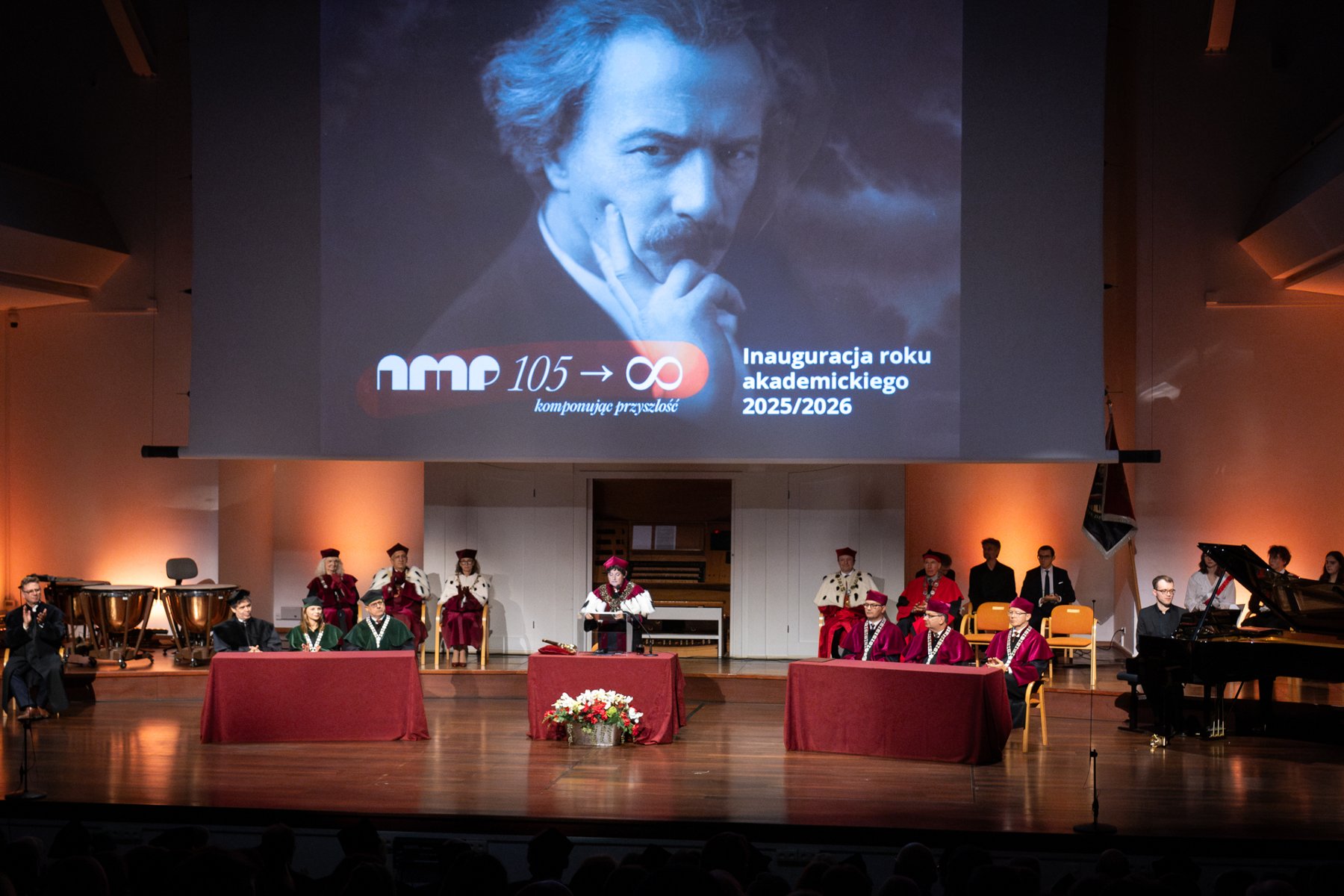 Zdjęcie przedstawia pracowników uczelni w togach z gronostajem za stołem. Nad nimi znajduje się plakat z wizerunkiem Paderwskiego.