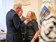 Zdjęcie przedstawia prezydenta przypinającego medal kobiecie. Obok stoi mężczyzna. Widać też flagę z herbem Poznania.