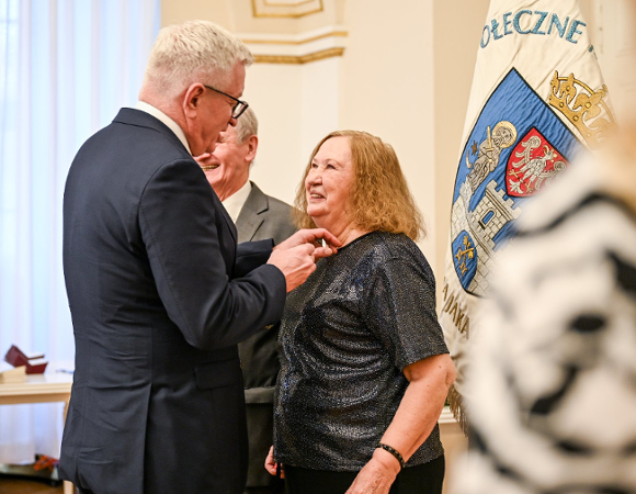 Zdjęcie przedstawia prezydenta przypinającego medal kobiecie. Obok stoi mężczyzna. Widać też flagę z herbem Poznania.