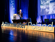 Zdjęcie przedstawia scenę. Przy mównicy stoi kobieta. Za jej plecami wyświetlane są zdjęcia zalanych ulic. Przed nią znajduje się napis Stormwater Poland.