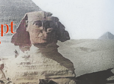 Zdjęcie przedstawia sfinksa. Przed nim na wielbładzie siedzi mężczyzna. W tle widać piramidę. Zdjęcie ma wyblakłe kolory.